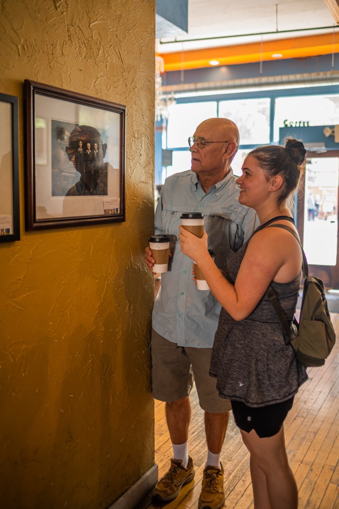 A man and a woman, holding coffee cups, look at art displayed on a yellow wall