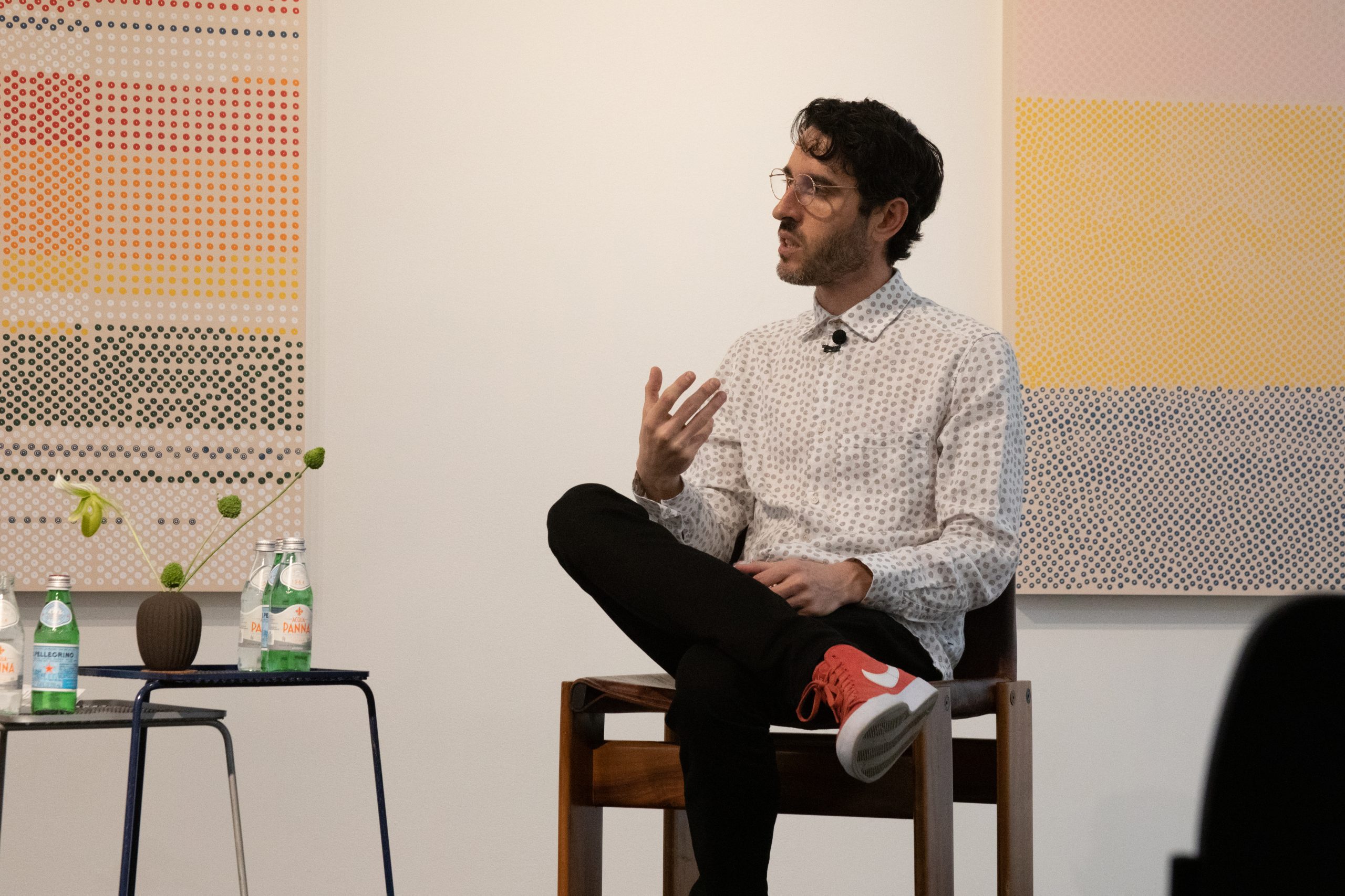 A seated man in a patterned shirt speaks to an audience in front of contemporary artwork