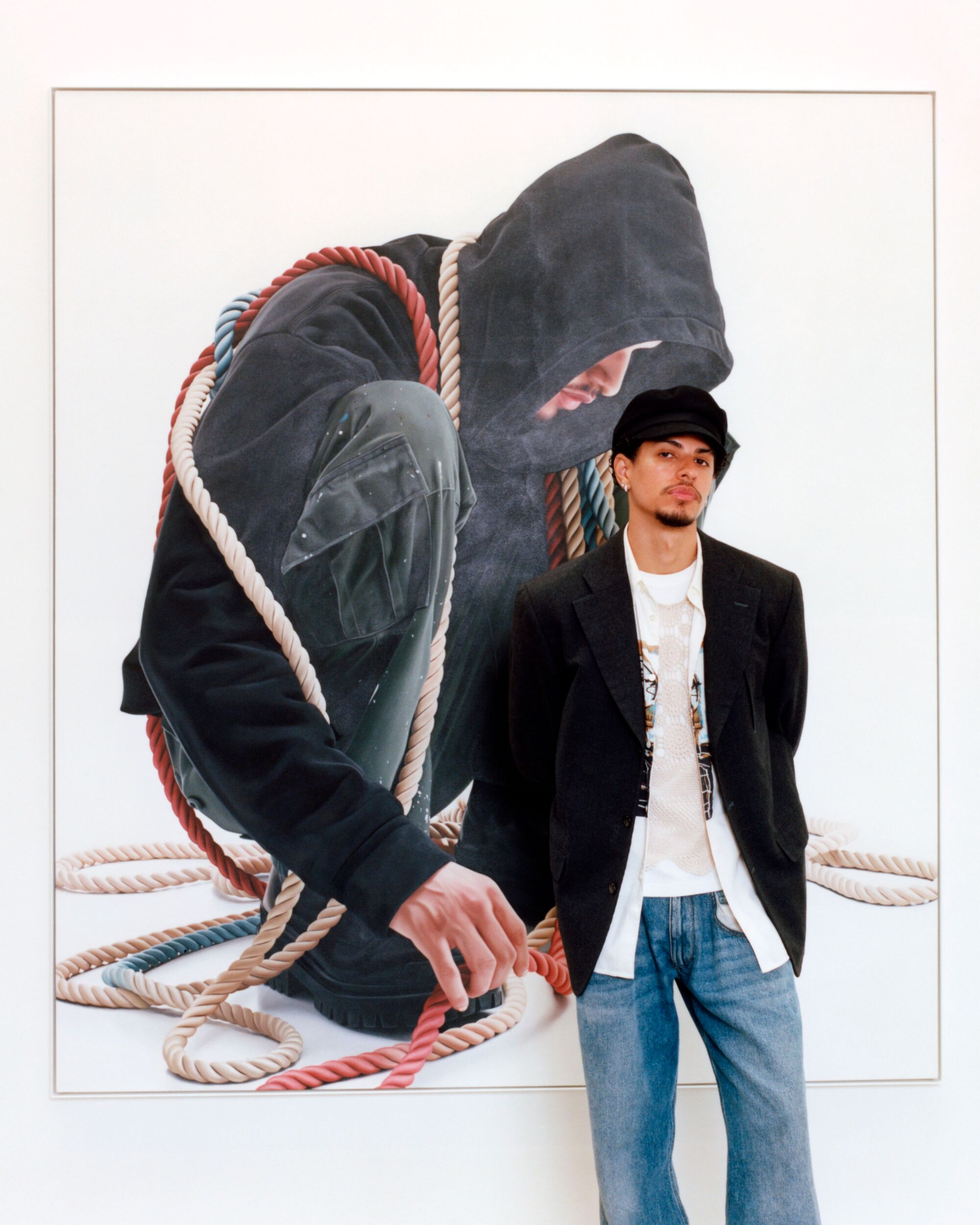 The artist stands in front of one of his paintings of a hooded figure draped in ropes.