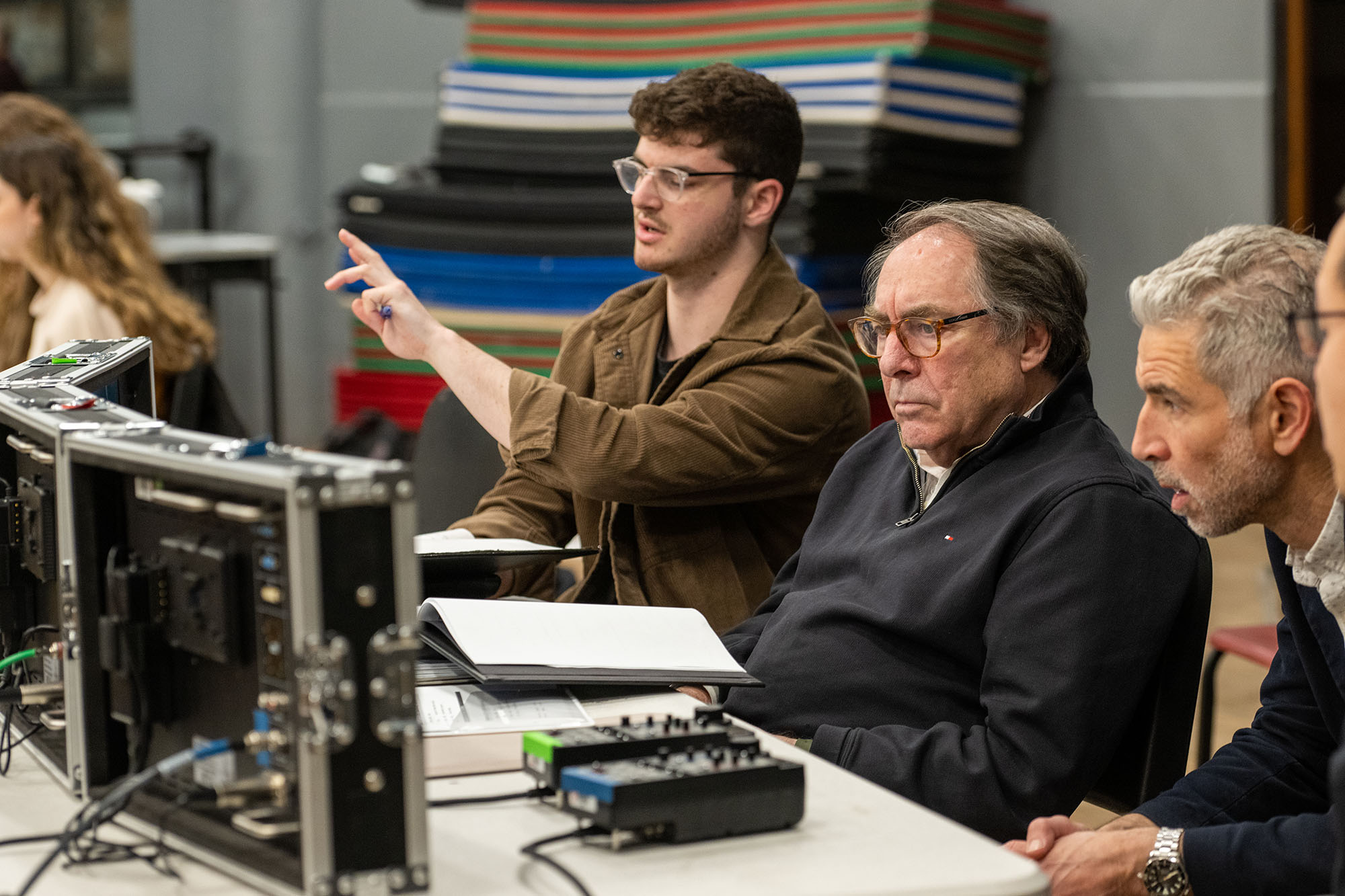 Photo: Director Eli Canter (left) and Paul Schneider, COM professor of film and television and department chair, discuss camera blocking during a rehearsal. A young man wearing glasses and a brown collared shirt points forward and speaks as an older man wearing glasses and a black collared shirt sits to his right. They both sit at a table with multiple mini screens in front of them.