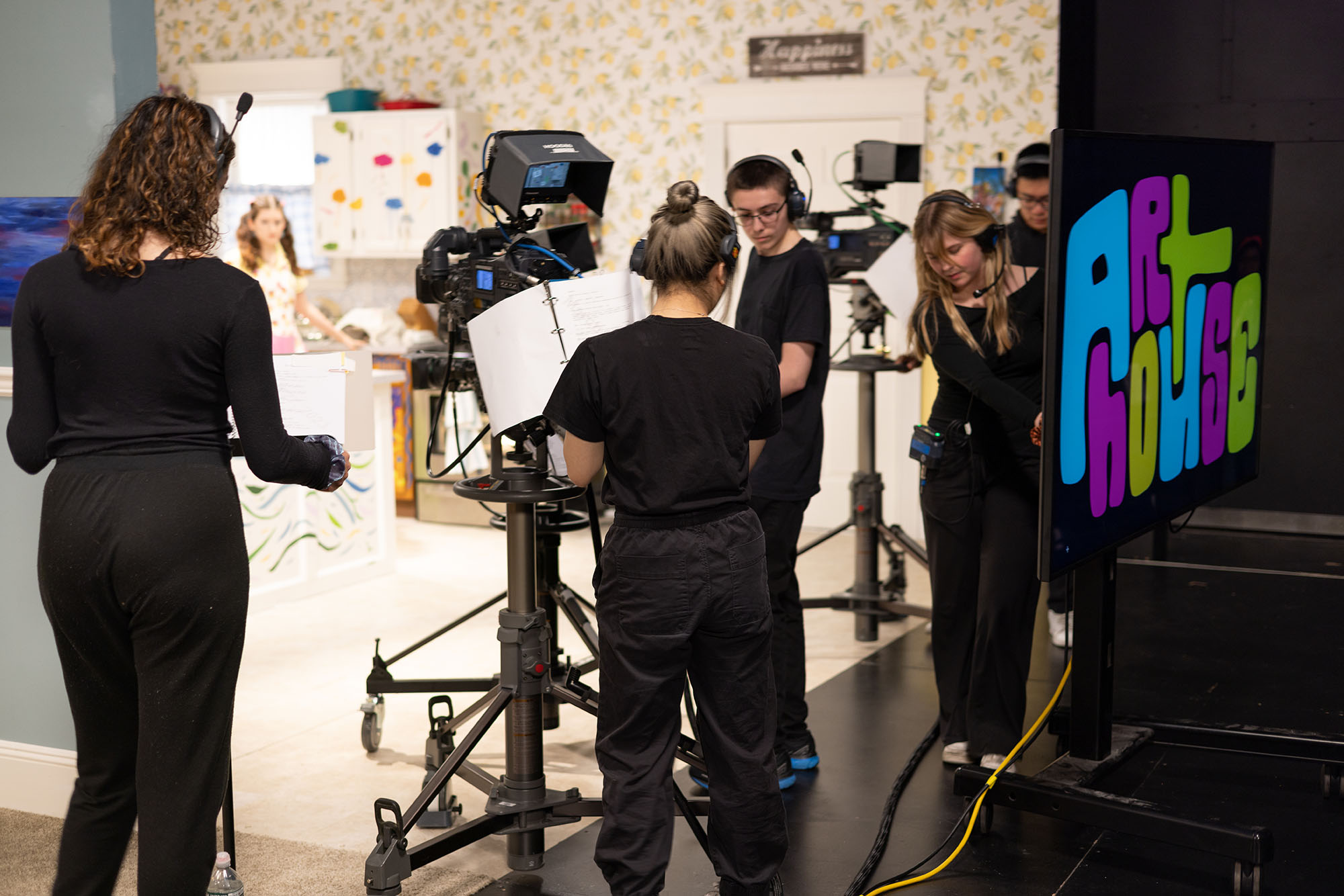 Photo: People wander and set up cameras and other lighting and recording equipment on the set of Art House. The various crew members wear all black outfits as they work and collaborate on the brightly lit, colorful set.
