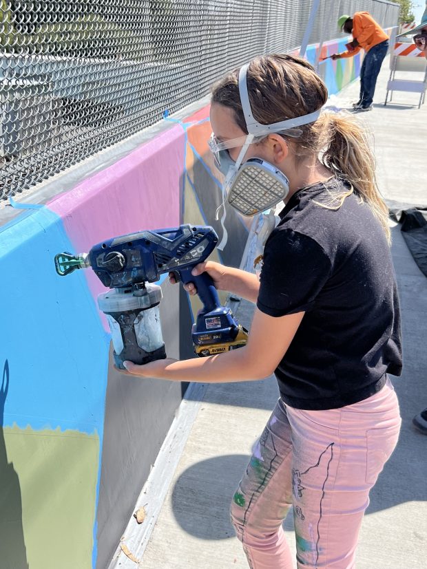Edin Goulart, 8, wears protective gear as she demonstrates her father's new paint sprayer on the Hawthorne Street mural. She worked on the mural with her dad, Ben Goulart, as part of the Eureka Street Art Festival, which wrapped up Saturday. (Heather Shelton/The Times-Standard)