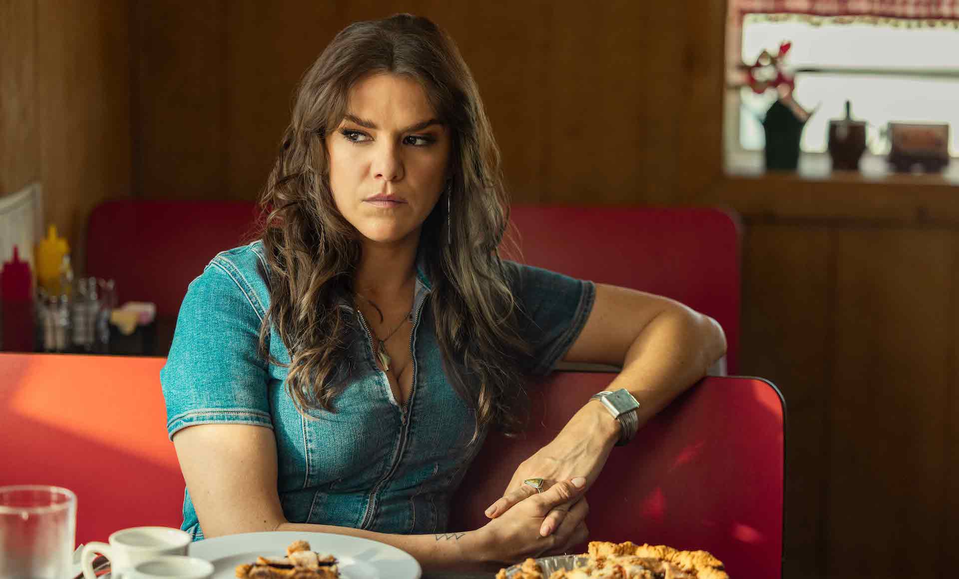 A woman with long, wavy brown hair wearing a denim zip-up top and sitting in a red diner booth in front of a whole pie; still from 