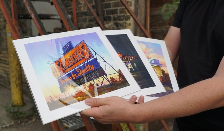 Person holds up art prints of an orange and blue sign that says Schneiders