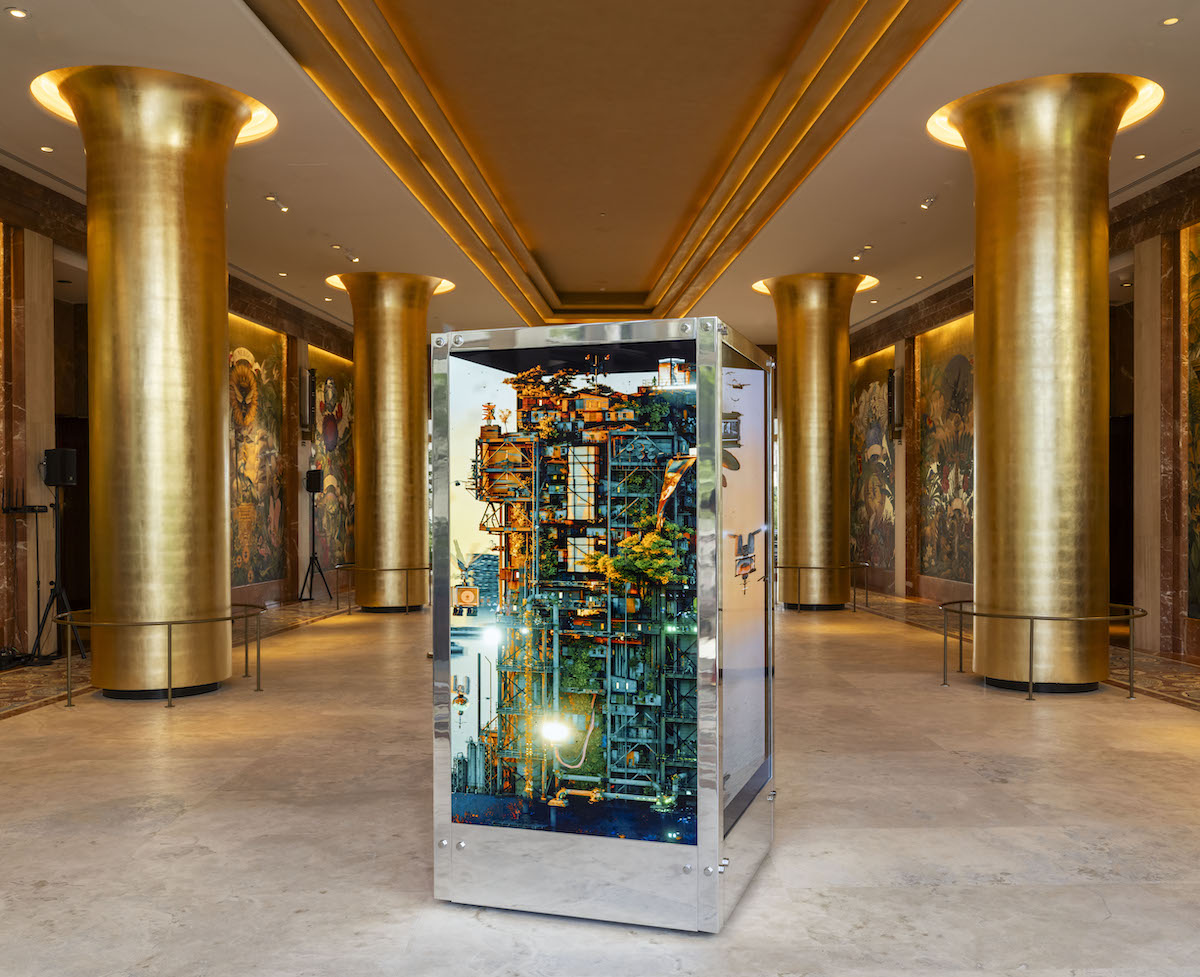 A six foot silver rectangular box with multicolored wires inside sits in a atrium of gold columns.