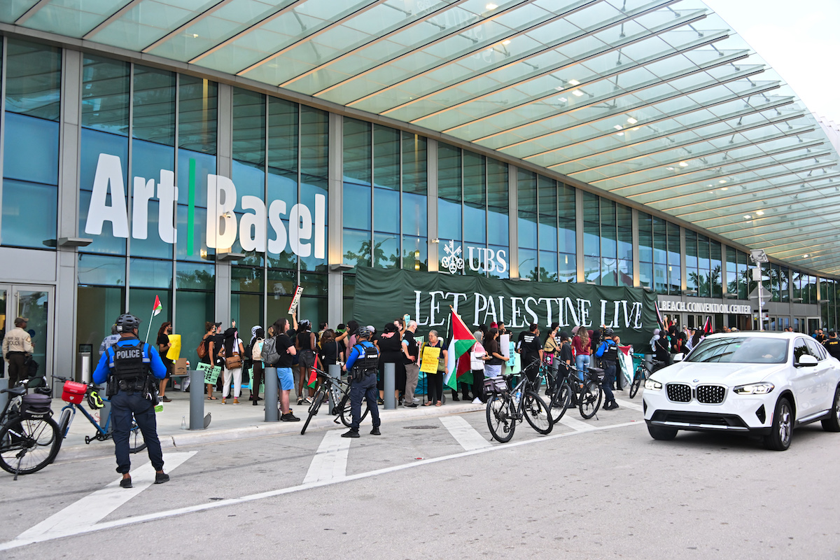 Protestors outside a convention center with the logo for Art Basel on it. Some hold a large banner that reads 'LET PALESTINE LIVE.'