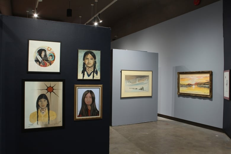 Four portraits of young women hang on a wall in the foreground and two landscape paintings on a wall in the background.