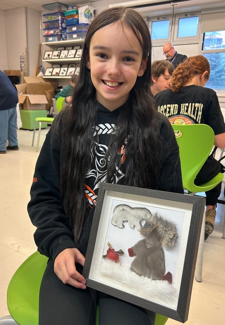 A girl in a classroom holding a shaddow box with a polar bear made from seal fur, and a figure of an inuit person wearing a seal jacket near a fire.