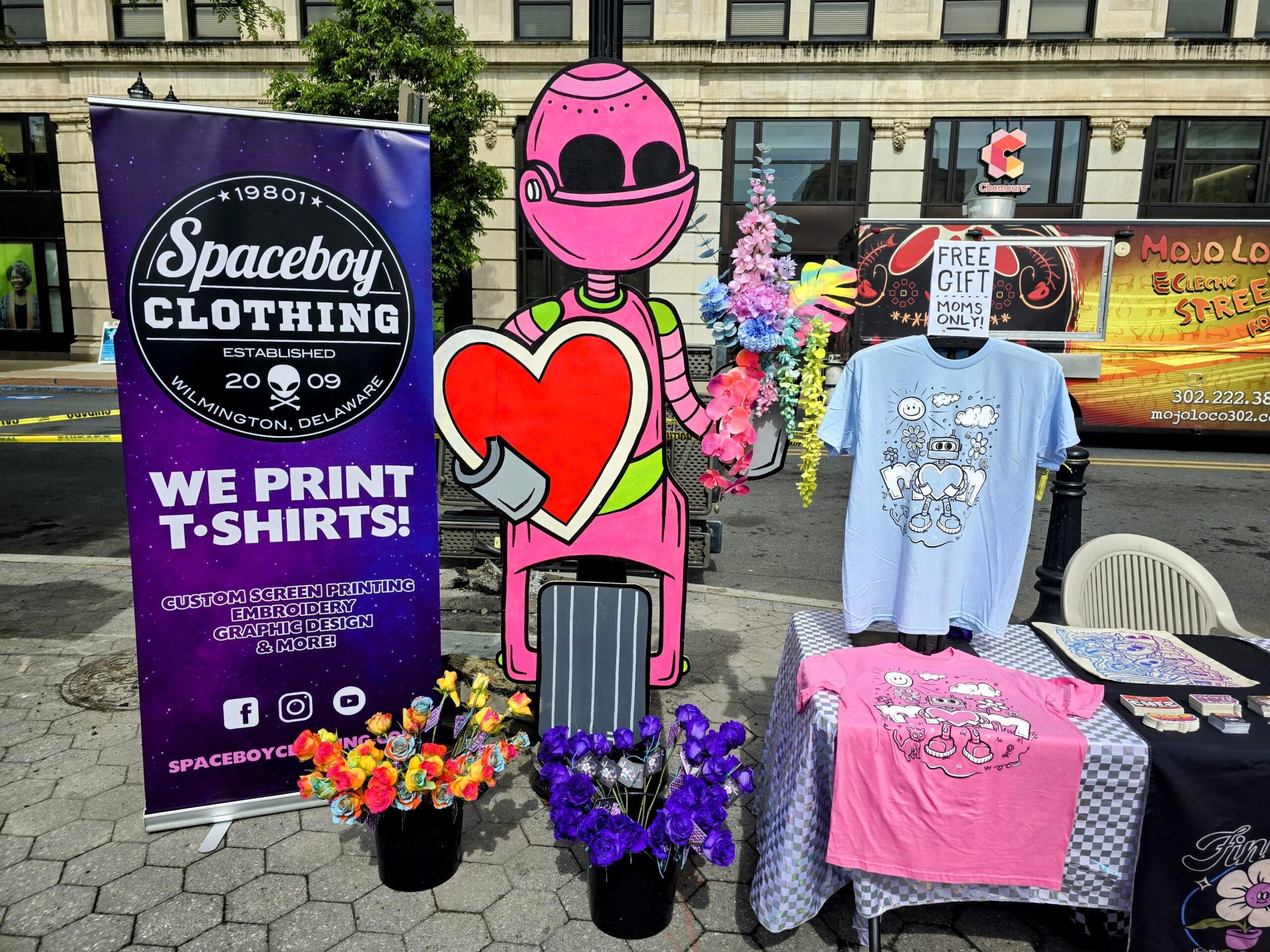 A street vendor booth with a banner for spaceboy clothing, featuring a large pink robot cutout, assorted t-shirts on display, and flower decorations.