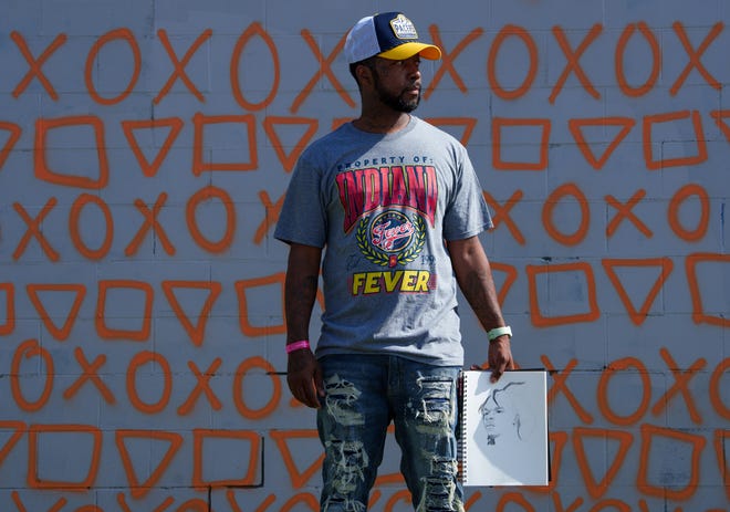 Indianapolis street artist Michael 'Kwazar' Martin poses Thursday, May 2, 2024, next to the wall where his next piece will be painted. In his hand is his sketchbook, and drawn is his next subject – Indianapolis Colts quarterback Anthony Richardson.